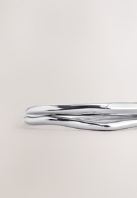 Metall-Haaraccessoire mit glatter, glänzender Chromoberfläche. Es zeichnet sich durch ein gedrehtes, fließendes Design aus und verjüngt sich an beiden Enden.