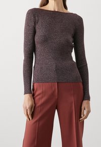  modell som bär en figurnära, djupröd, långärmad topp som är matchad med högmidjade, löst sittande vinröda byxor framför en neutral bakgrund.