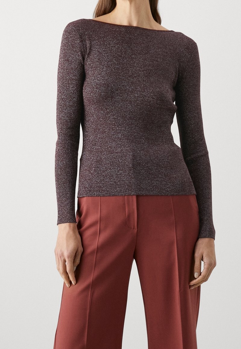 modell som bär en figurnära, djupröd, långärmad topp som är matchad med högmidjade, löst sittande vinröda byxor framför en neutral bakgrund.