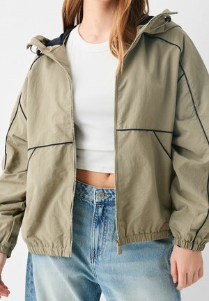Ung person iført en beige hættejakke med lynlås over en hvid cropped top og lyseblå højtaljede jeans.