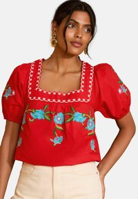 Blusa rossa in cotone con maniche a palloncino, scollatura quadrata e ricami floreali blu. Una cucitura bianca a contrasto delimita la scollatura.