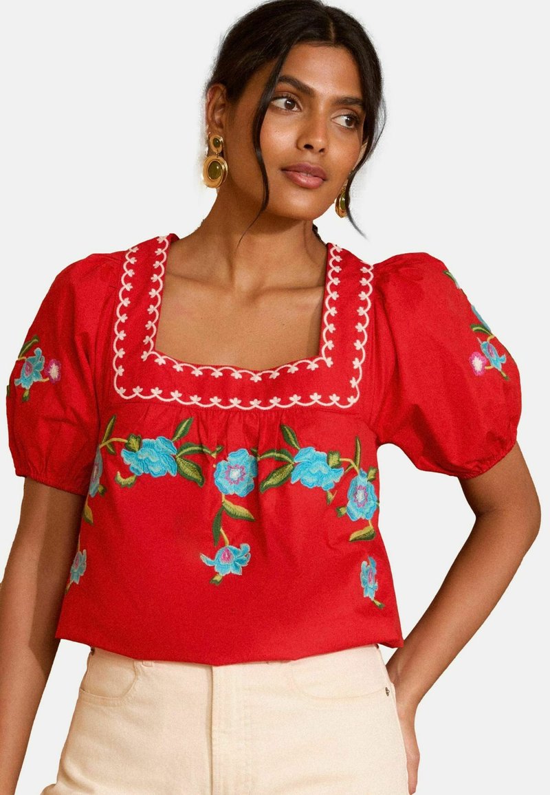 Blusa rossa in cotone con maniche a palloncino, scollatura quadrata e ricami floreali blu. Una cucitura bianca a contrasto delimita la scollatura.