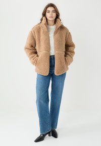 Beżowa kurtka z futerkiem ekologiczny o wysokim kołnierzu, zapinana na zamek, z dwoma przednimi kieszeniami, noszona na jasnym swetrze i niebieskich dżinsach.