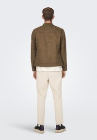 Jachetă din suede maro cu un design aerodinamic, pantaloni bej și pantofi negri cu accente mov. Stil minimalist, fără logo-uri vizibile.