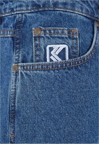 Vaqueros de mezclilla azul oscuro con un bolsillo trasero con un logo bordado y remaches de color cobre. La tela tiene un acabado ligeramente texturizado.