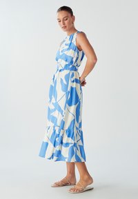 Kobieta ubrana w bezrękawową midi sukienkę w niebiesko-białe abstrakcyjne wzory z falbaniastym dołem oraz płaskie beżowe sandały, stojąca z rękami za plecami.