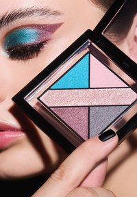 Primo piano di una persona con ombretto turchese e viola che tiene una palette di ombretti con tonalità blu, rosa, lavanda, argento e oro rosa.