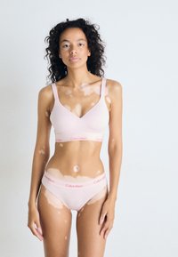 Lichtroze Calvin Klein bralette met een V-hals en brede banden, gecombineerd met bijpassende slips met een logo tailleband. Soepele textuur, minimalistisch ontwerp.