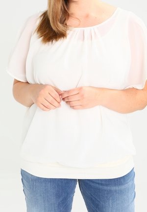 Vrouw met lichtbruin haar, gekleed in een losse witte blouse en blauwe spijkerbroek, handen licht geklemd voor zich.