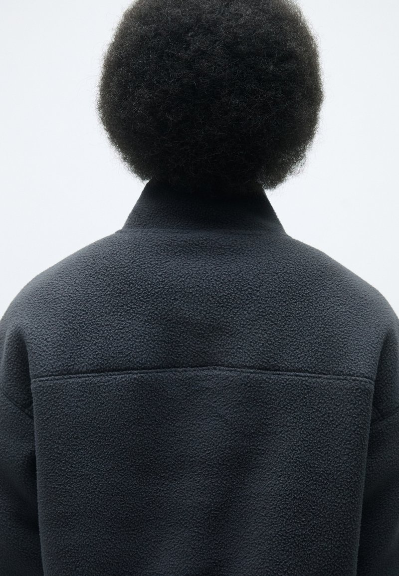 Mörkgrå fleecejacka med mjuk textur, hög krage och minimalistisk design. Baksidan visar en sömdetalj över axlarna.