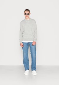 Sweatshirt cinza com gola redonda, camiseta branca por baixo, jeans azuis e ténis brancos. Acessórios com óculos de sol escuros. Modelo em pé.