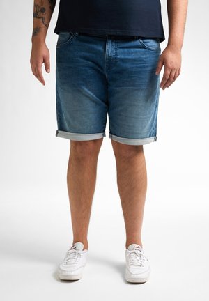 Denimshorts i mørkeblå med et falmet mønster. Rullekant og to frontlommer. Båret med hvide sneakers på en lys baggrund.