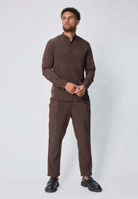 Chemise marron à manches longues avec un col mandarin, fermeture à boutons sur le devant, et pantalon ajusté assorti, accompagné de chaussures noires. Texture du tissu lisse.