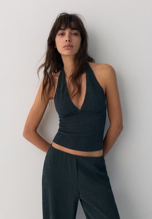 Femme debout contre un mur blanc, portant un haut à col halter texturé foncé et un pantalon large assorti, mains derrière le dos, expression neutre.