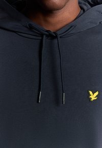 Felpa con cappuccio della marina, realizzata in tessuto testurizzato, con lacci neri e un logo ad aquila ricamato giallo sul petto.