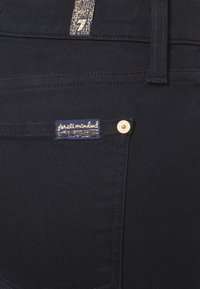 Tylnia kieszeń ciemnych dżinsów z niebieską metką materiałową z napisem „for all mankind” oraz miedzianym ćwiekiem na rogu kieszeni.