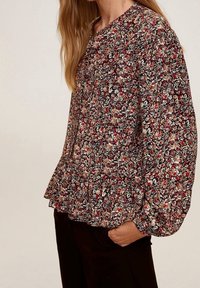 Floral blouse in zwart, met kleine rode, witte en gele bloemen. Heeft een geplooide zoom, ronde halslijn en lange pofmouwen.
