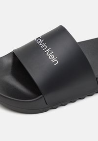 Infradito nera con una vasta fascia opaca che presenta il logo "Calvin Klein" in rilievo. Suola testurizzata con scanalature per una buona aderenza. Design casual.
