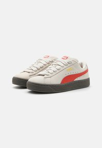 Sneaker in suede beige con striscia laterale rossa, suola in gomma texturizzata grigia e design con lacci. Logo del marchio giallo sul lato esterno.