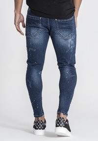 Vaqueros de denim oscuros de corte ajustado con detalles desgastados, arrugas y bolsillos traseros. Combinados con zapatillas negras con patrón.