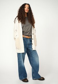 Cárdigan crema de gran tamaño con acentos texturizados, camiseta gris, jeans de pierna ancha azules y botas marrones. Fondo liso.