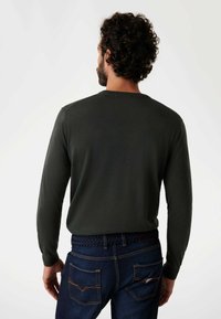 Dunkelgrüner Strickpullover mit langen Ärmeln und rundem Ausschnitt, getragen mit blauen Jeans, die einen geflochtenen Gürtel und sichtbare Details an den Gesäßtaschen aufweisen.