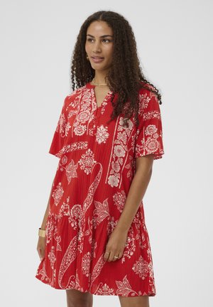 Kobieta z kręconymi włosami, ubrana w czerwoną sukienkę w kwiatowy wzór z krótkim rękawem, stoi na jednolitym tle, lekko patrząc na bok.