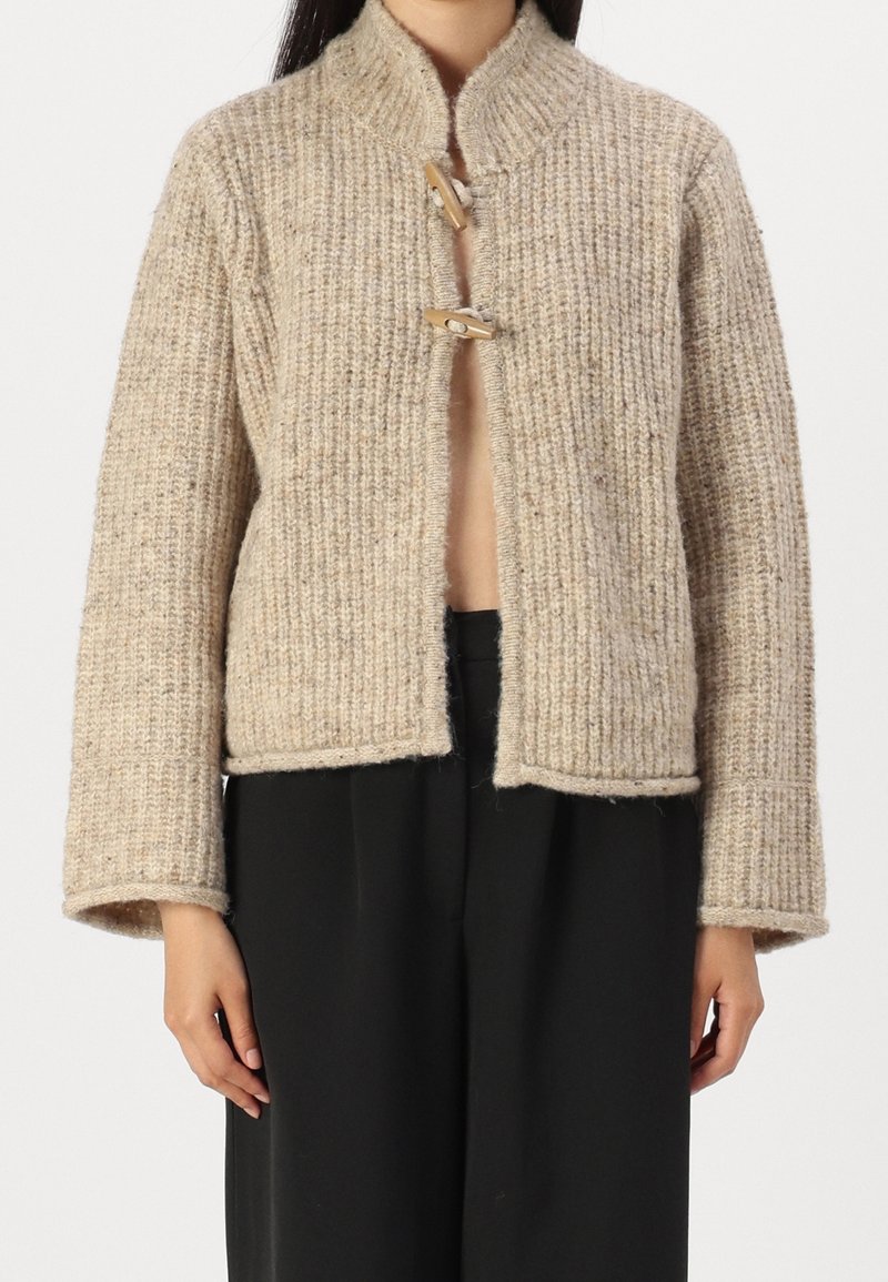 Cardigan beige en maille avec une texture épaisse et deux fermetures à bascule en bois, porté sur un pantalon noir à jambes larges.
