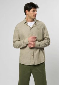 Lichtbeige overhemd met knopen en een textuur, voorzien van een subtiele logo op de borst, gecombineerd met olijfgroene broeken.
