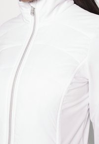 Biała pikowana kurtka z zamkiem błyskawicznym z przodu i wysokim kołnierzem, noszona przez osobę, ukazująca szczegóły górnej części torsu i ramienia.