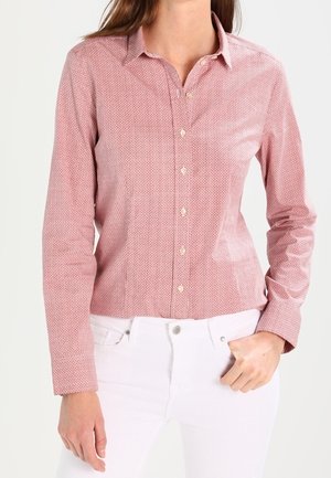 Mujer con una camisa de botones rosa de manga larga con estampado y cuello, combinada con pantalones blancos, de pie con una mano en el bolsillo.