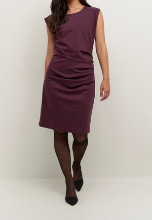 Femme portant une robe prune sans manches longueur genou avec des détails froncés, des collants noirs transparents et des escarpins noirs à bouts pointus, debout devant un fond uni.