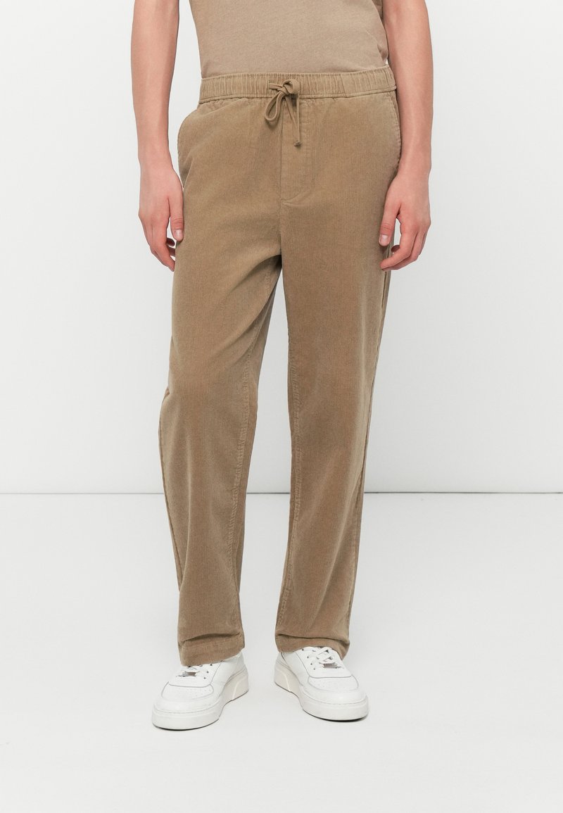 Tan corduroy pants with an elastic waistband and drawstring, featuring side pockets and a relaxed fit, paired with white sneakers.