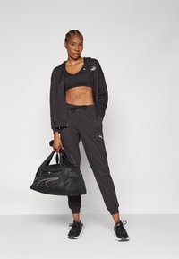 Abbigliamento sportivo nero composto da un top corto, una felpa con zip e pantaloni sportivi. Gli accessori includono una borsa da palestra nera e sneakers nere.
