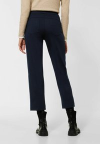 Marineblaue Hose mit elastischem Bund und schmal zulaufendem Bein, ausgestattet mit zwei Gesäßtaschen und einer glatten, weichen Textur. Tragen mit schwarzen Knöchelstiefeln.