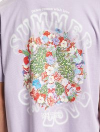 Lavender short-sleeve t-shirt featuring a floral design with red, blue, and white flowers, surrounded by text "SUMMER" and "LOVE COMES WITH PEACE."