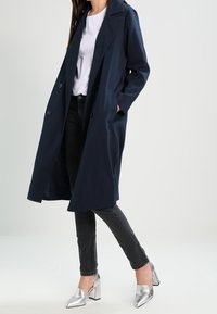 Marinblå lång kappa med krage, två framfickor, knappstängning, tillsammans med svarta jeans och silverfärgade högklackade skor. Slät textur.