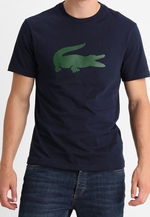 Homme portant un t-shirt bleu marine avec un grand motif crocodile vert sur la poitrine et un jean bleu, debout, bras détendus.