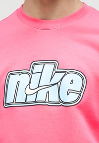 Κοντινό πλάνο ενός ατόμου που φορά ροζ πουκάμισο με μεγάλο λευκό και μαύρο λογότυπο Nike στο κέντρο του στήθους.