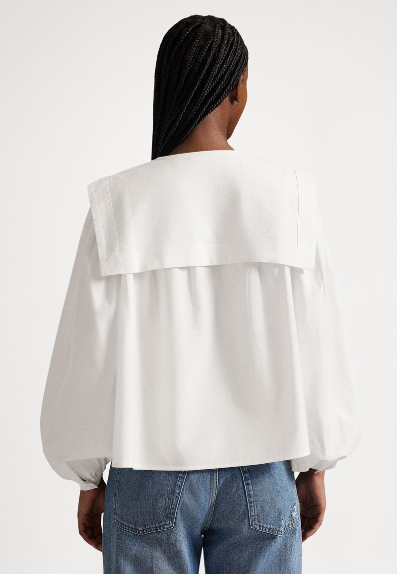 Persoon met lang gevlochten haar draagt een losse witte blouse met een grote matrozenkraag en blauwe spijkerbroek, kijkt weg van de camera.