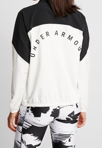 Chaqueta deportiva en blanco y negro con cintura elástica, que presenta un logo en una fuente negra en negrita en la parte posterior y una textura de tela suave y elegante.