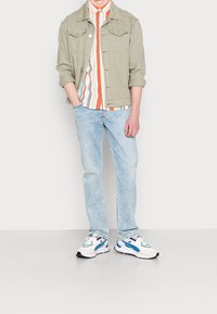 Licht olijfgroen denim jack, gestreept shirt met oranje en blauwe accenten, lichtblauwe spijkerbroek en veelkleurige sportieve sneakers.