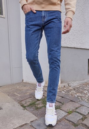 Osoba v modrých úzkých džínách, béžové mikině a bílých teniskách Adidas kráčí po dlážděném chodníku vedle zdi budovy.