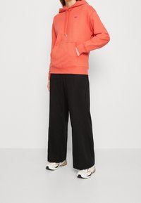 Osoba ubrana w koralowy hoodie, czarne szerokie spodnie i białe sneakersy stoi z jedną ręką w kieszeni hoodie'u na tle jednolitym.