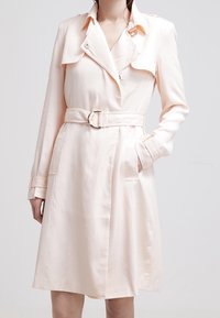 Jasnoróżowy płaszcz typu trencz z podwójnym zapięciem, paskiem w talii i bocznymi kieszeniami. Wykonany z gładkiej tkaniny, z kołnierzem wciętym.