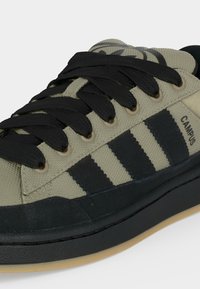 Μπεζ και μαύρο αθλητικό παπούτσι Adidas Campus με μαύρα κορδόνια, τρεις μαύρες ρίγες και μπεζ καουτσούκ σόλα σε λευκό φόντο.