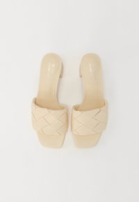 Beige slip-on sandals with a woven strap design and a square toe. Smooth leather texture and a geometric pattern accent the upper.