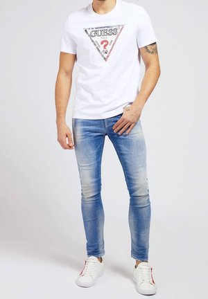 White T-shirt with a triangular "GUESS?" graphic in black, red, and blue, paired with light blue skinny jeans and white sneakers.