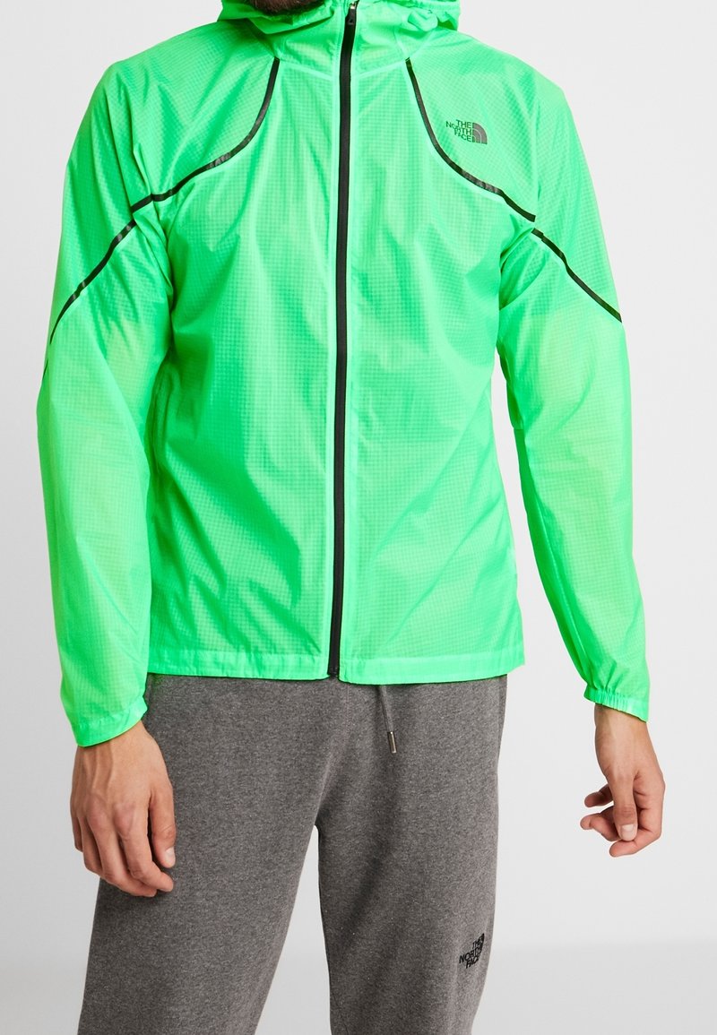 Homme portant une veste à capuche zippée vert vif et un pantalon de survêtement gris, tous deux avec le logo The North Face.