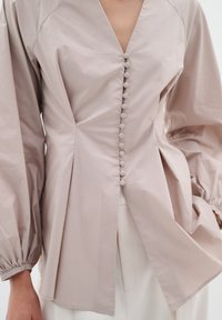 Camicia abbottonata in morbido tessuto beige con scollo a V, design plissettato e maniche lunghe a sbuffo. Presenta bottoni tondi a intervalli regolari lungo la parte anteriore.